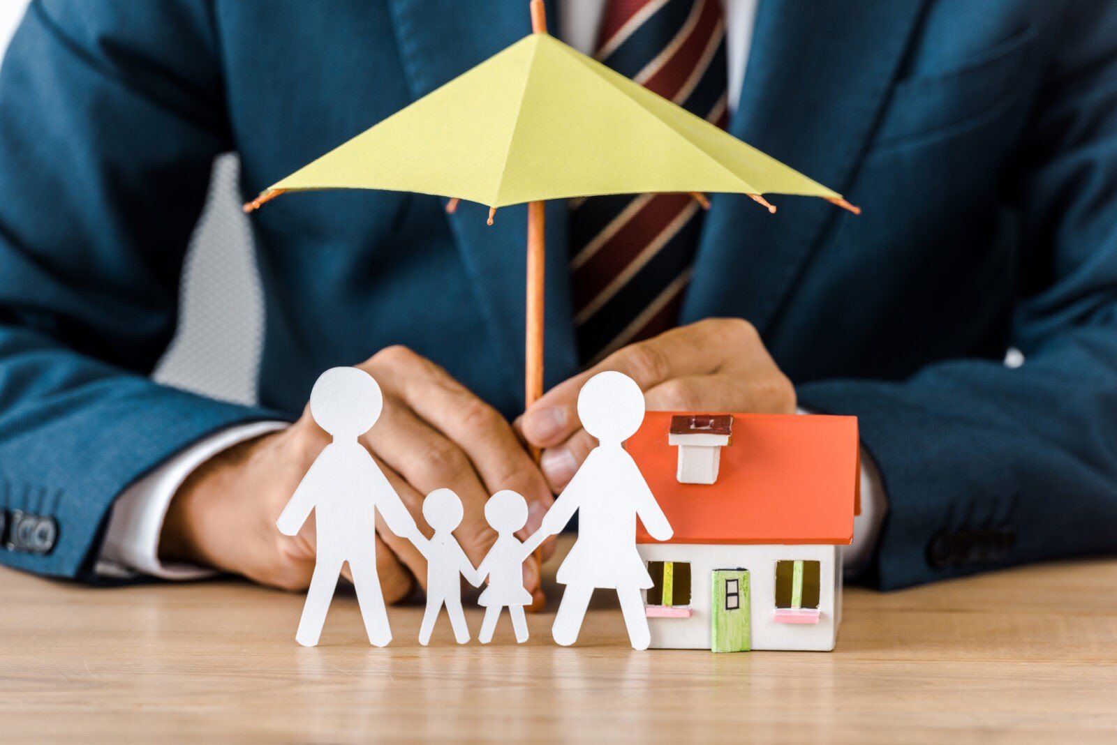 Male hands with paper cut family, house model and umbrella on wooden table, life insurance